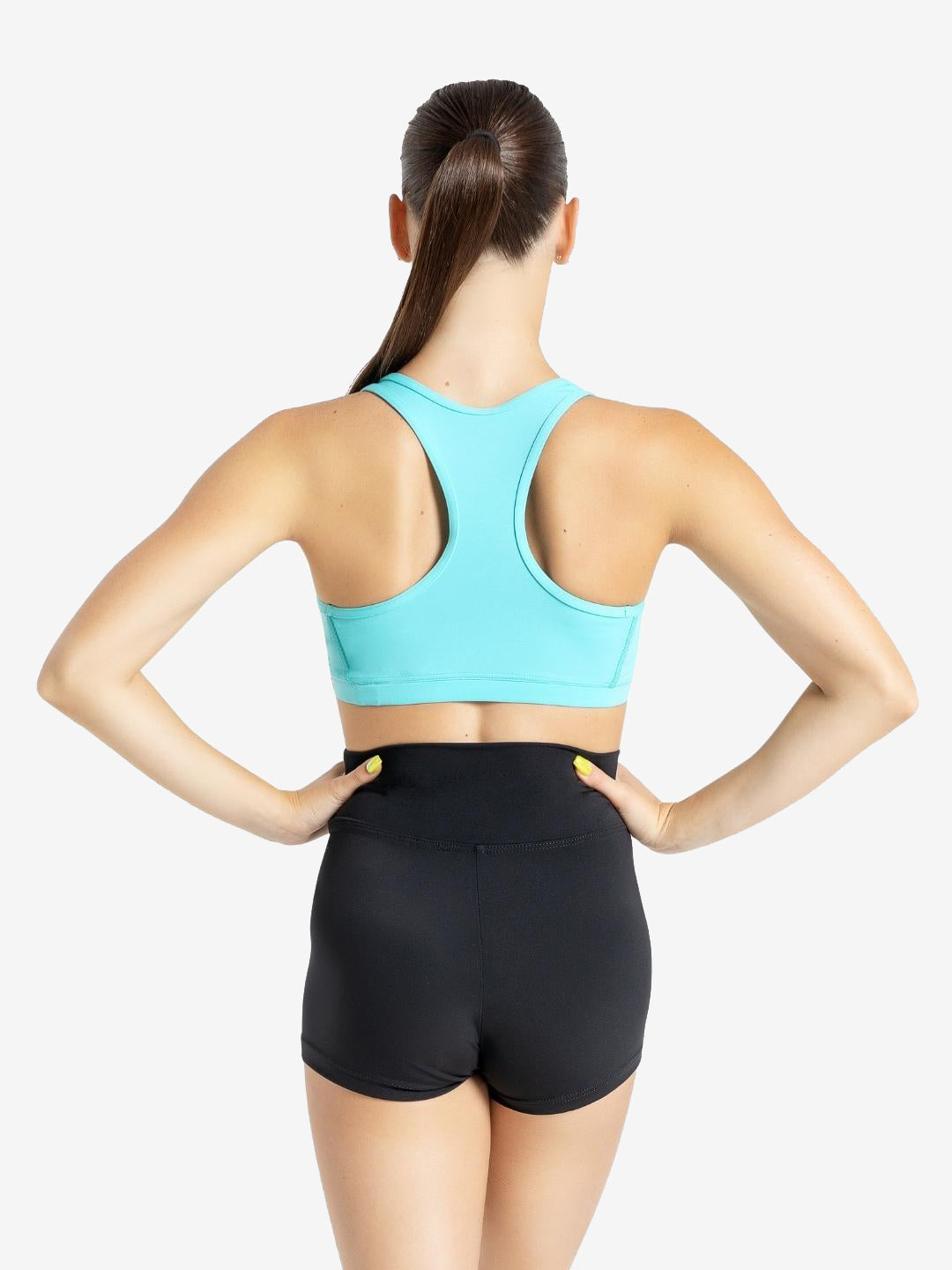 The back of a woman with a long brown ponytail draped over her front shoulder wearing a turquoise sports bra and high waisted black shorts. Her hands are on her hips with painted yellow nails.