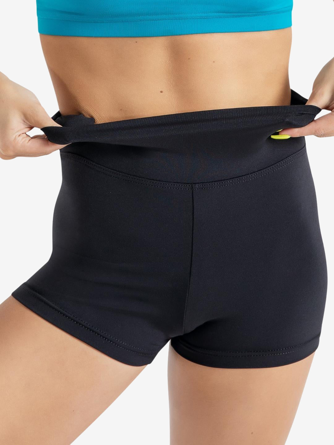 A close-up of a person wearing a blue sports top and black high-waisted athletic shorts. She is adjusting the waistband of the shorts, pulling it up slightly with both hands. The background is plain light grey.