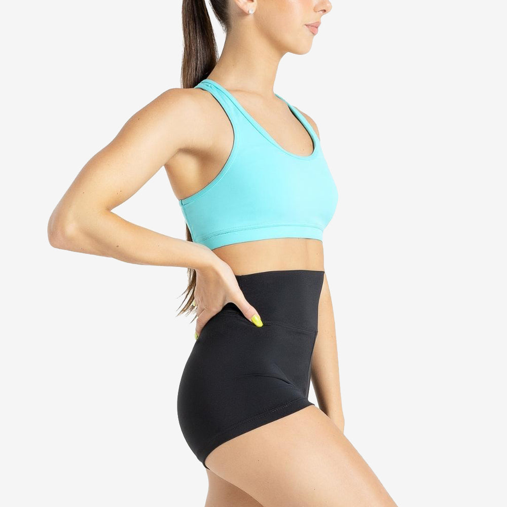 
                      
                        A woman with long brown hair in a ponytail is wearing an aqua colored sports bra and high-waisted black athletic shorts. She is standing in profile with one hand on her hip and looking forward. The background is light grey.
                      
                    