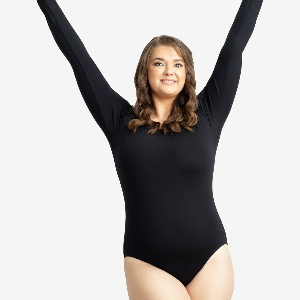 A woman with wavy brown hair is smiling and stretching her arms upwards. She is wearing a long-sleeved black bodysuit and posing confidently against a plain white background.