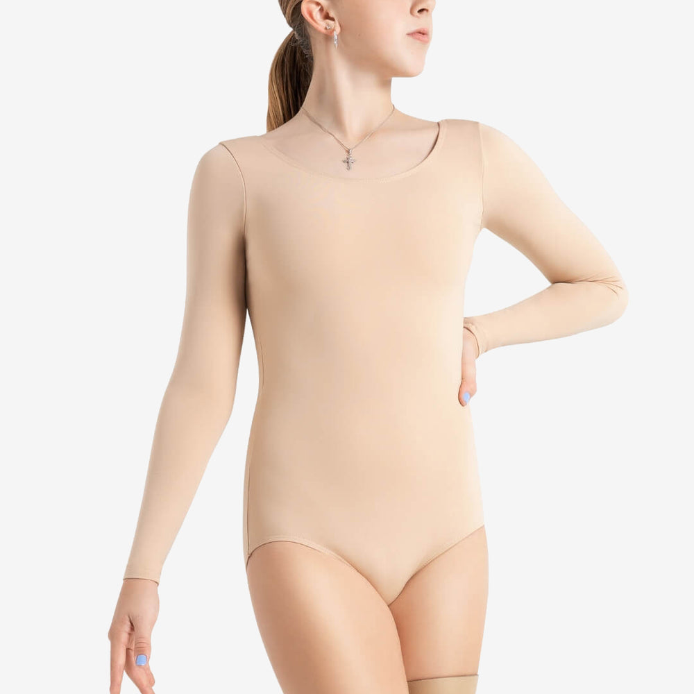 A young woman with her hair pulled back is wearing a beige long-sleeve leotard. She is looking to her left with her right arm on her hip and her left arm bent. She wears a small necklace with a cross pendant, and a neutral background highlights her attire.
