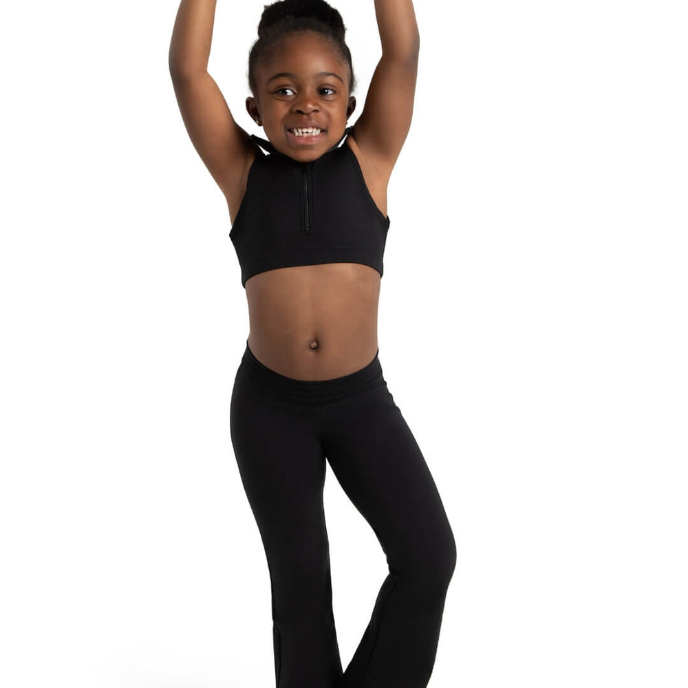
                      
                        A young child smiles joyfully while standing in a ballet pose with arms raised overhead. They are wearing a black crop top and matching black pants. The background is plain white.
                      
                    