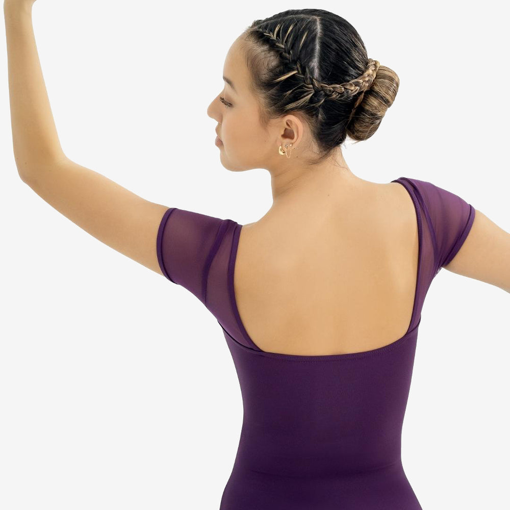 A ballerina in a purple eggplant tone leotard showcases the back square cut mesh sleeved design. Left arm extended up and to the side, right arm down and to the side. Her hair is braided and pulled back into a bun.