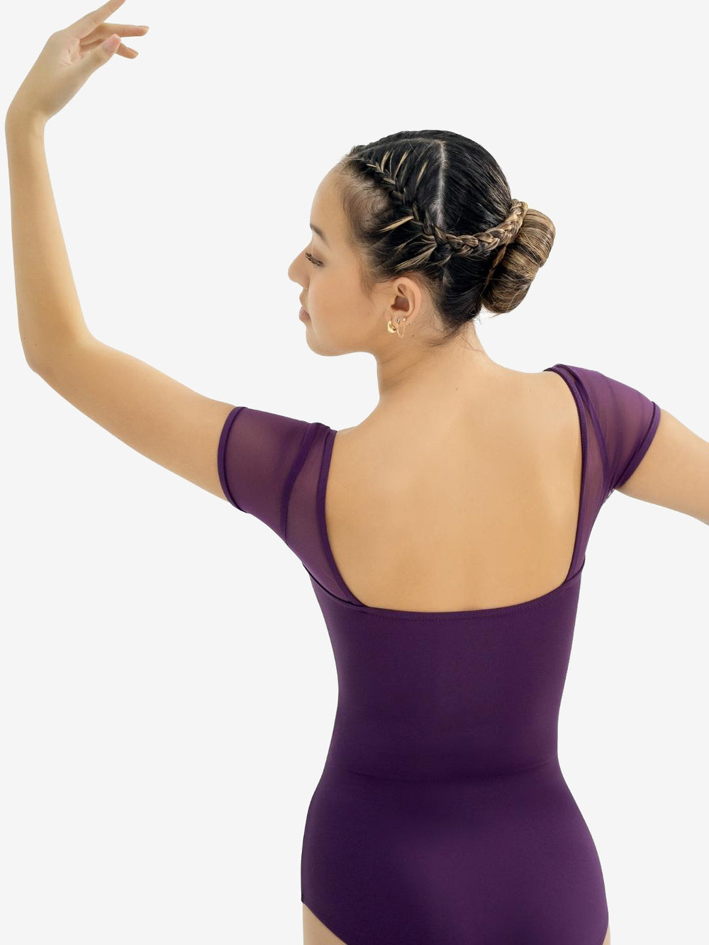 A ballerina in a purple eggplant tone leotard showcases the back square cut mesh sleeved design. Left arm extended up and to the side, right arm down and to the side. Her hair is braided and pulled back into a bun.