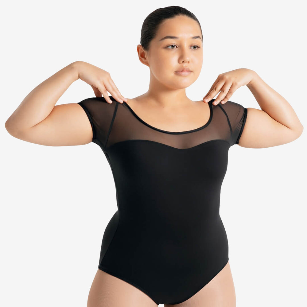 
                      
                        A female with slicked back black hair, stands with her hands on her shoulders, wearing a mesh short sleeve upper black sweetheart neckline leotard and skintone tights.
                      
                    