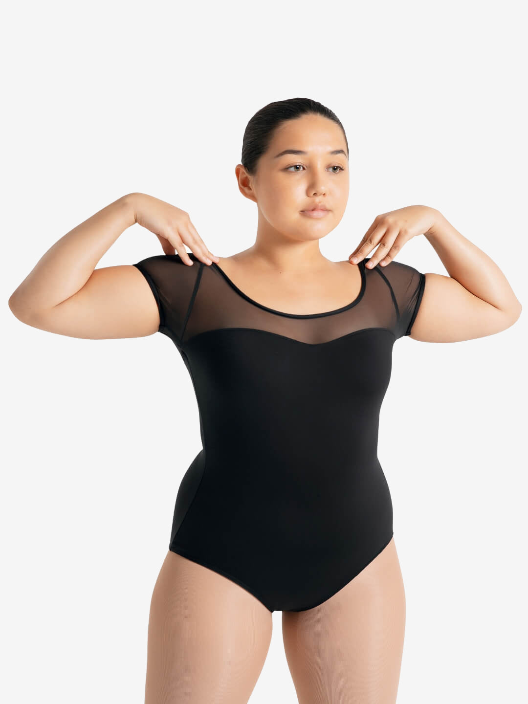 A female with slicked back black hair, stands with her hands on her shoulders, wearing a mesh short sleeve upper black sweetheart neckline leotard and skintone tights.