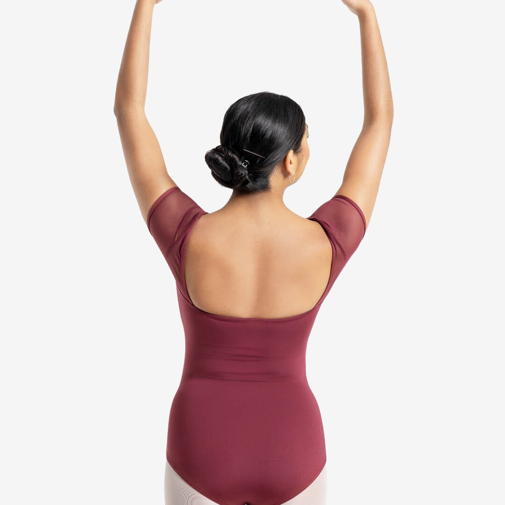 
                      
                        A dancer lifts her hands with yellow painted nails, gracefully over her head. Her black hair bobby-pinned and twisted into a bun. Showcasing the back square cut design of a short-mesh-sleeve burgundy leotard.
                      
                    