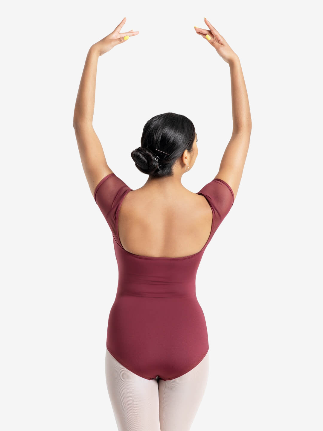 A dancer lifts her hands with yellow painted nails, gracefully over her head. Her black hair bobby-pinned and twisted into a bun. Showcasing the back square cut design of a short-mesh-sleeve burgundy leotard.
