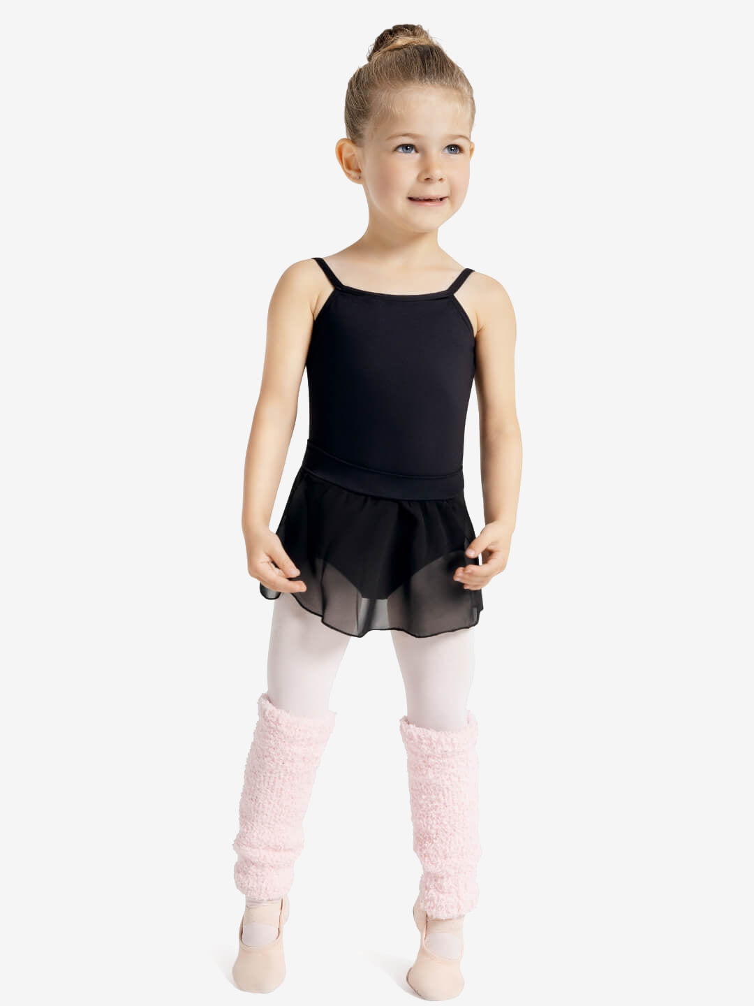 A young child with a blonde top ballet bun smiles off to the side and holds arms in a ballet position in front of her slightly. She is wearing a black leotard and pull-on black ballet skirt, ballet pink tights, pink legwarmers, and ballet shoes, against a light grey solid background.