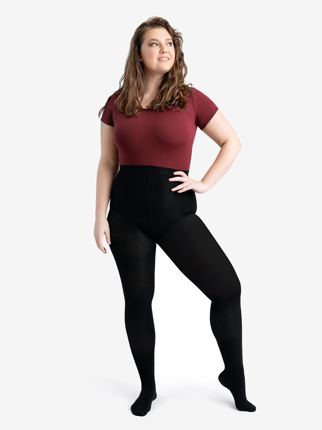 A woman stands with her hand on her hip, she has wavy hair and looks up and to the side. She is wearing a burgundy, short sleeve leotard and black tights on top.