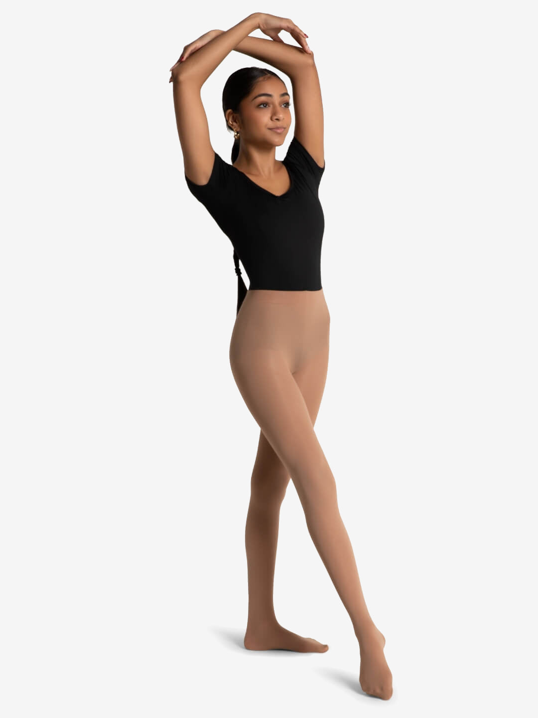 A young woman stands with her weight on her back leg and front leg extended and pointed forward. Her hand are over her head. She is wearing a black short sleeve leotard and tan tights, her long black hair is seen tied behind and flowing down her back. Plain background.