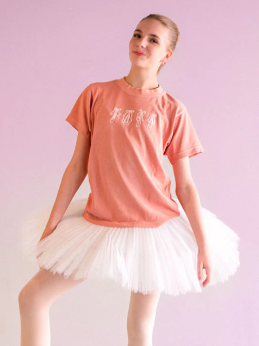 Girl wearing a pink t-shirt with pointe shoe graphic and a white tutu against a light pink background