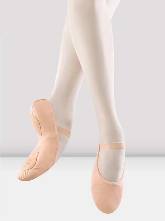 A close-up of a pair of legs in pink ballet shoes, pointing down toward the ground. The back foot is turned to show the split-sole bottom. Light grey background.