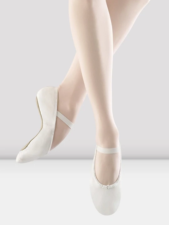 A close-up of a person's legs wearing white, leather ballet shoes with a single elastic strap across the top of the foot. The feet are pointing down as if mid-air, the back leg bent to show the flexibility of the shoe and pointe of the foot.