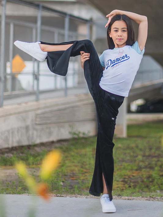 A young girl stands outside on one foot, the other leg lifted in the air and held by her hand. Her opposite hand is over her head. She has long brown hair, black pants that have open slits from the knee to the ankle and she wears a raglan style white and light blue tee with blue text that reads DANCER