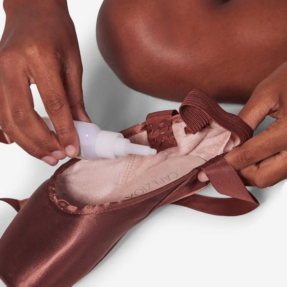
                      
                        Person applying a product to a brown pointe shoe on a plain background
                      
                    