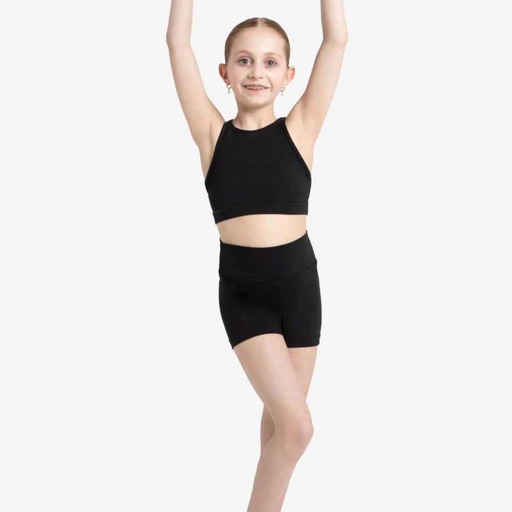 
                      
                        A young girl with blonde hair pulled into a bun, stands with arms overhead, wearing a black crop top and high waisted dance shorts
                      
                    